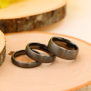 May include: Three black matte finish wedding bands on a wooden surface. The bands are stacked on top of each other.