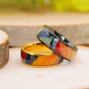 May include: Two rings with colorful wood grain in shades of blue, green, red, and orange. One ring has a gold interior and a black exterior band. The other ring has a gold interior and a colorful exterior band.