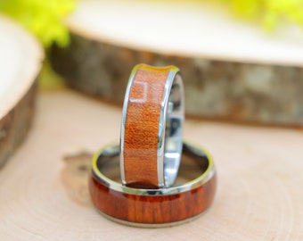 May include: Two rings with a polished wood inlay and a silver-colored metal band. The wood has a reddish-brown hue. The rings are displayed on a light-colored wooden surface.