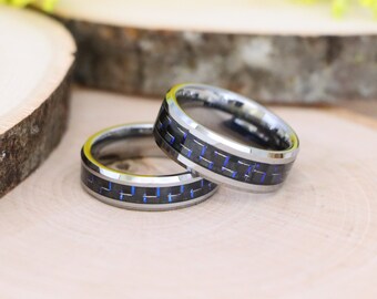 May include: Two silver rings with black and blue carbon fiber inlays. The rings are displayed on a light wood surface. The rings have a polished finish and a modern design, suitable for a wedding or anniversary.