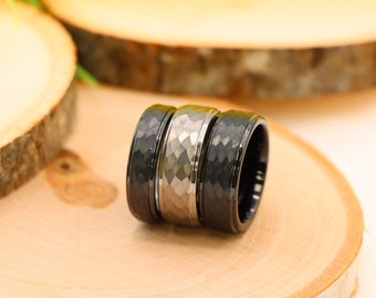 May include: Three rings with textured surfaces are displayed on a wooden slice. The rings have a hammered metal design. One ring is silver, and the other two are black. The rings are a jewelry item.