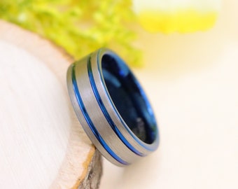May include: A silver-colored ring with two thin blue stripes and a blue interior. The ring is set on a piece of wood, with a blurred background of yellow and green.