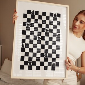 May include: Black and white framed artwork featuring a chess board design. The artwork has a light wood frame and a black and white checkered pattern. The chess pieces are represented by silhouettes.