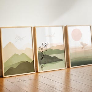 May include: Three framed art prints with a minimalist landscape theme. Each print features a different watercolor scene with mountains, trees, and a sun. The color palette includes shades of green, beige, and pink. The frames are made of light-colored wood.