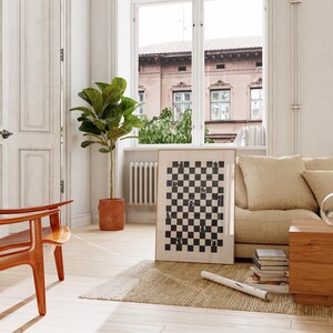 May include: A framed black and white chess board print leans against a beige sofa in a well-lit room. A wooden chair and a potted plant are in the foreground. A window with a view of a building is in the background.