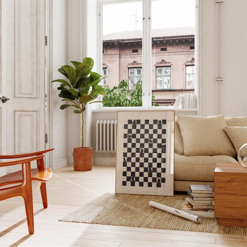 May include: A framed black and white chess board print leans against a beige sofa in a well-lit room. A wooden chair and a potted plant are in the foreground. A window with a view of a building is in the background.