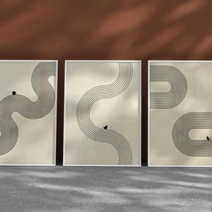 May include: Set of three framed abstract art prints. Each print features a beige background with a black, curved linear design. The frames are white, and the artwork is displayed against a brown wall and gray floor.