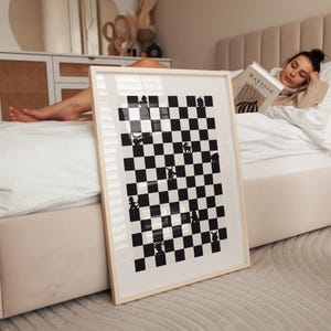 May include: A framed black and white art print featuring a checkerboard design with whimsical silhouette figures. The artwork is displayed in a light-colored wooden frame. The print is leaning against a bed with a person reading a book.
