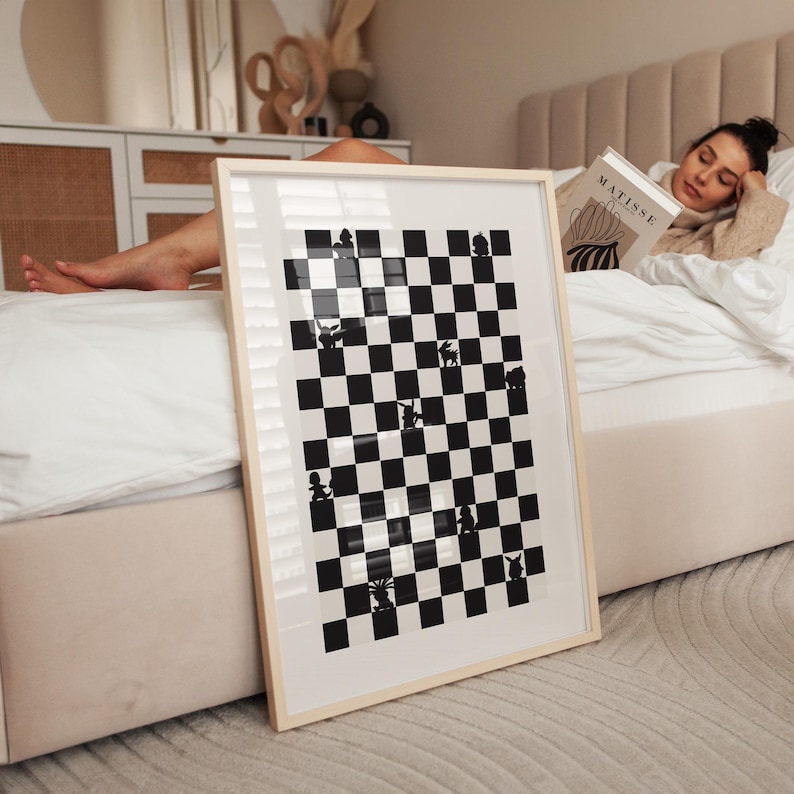 May include: A framed black and white art print featuring a checkerboard design with whimsical silhouette figures. The artwork is displayed in a light-colored wooden frame. The print is leaning against a bed with a person reading a book.