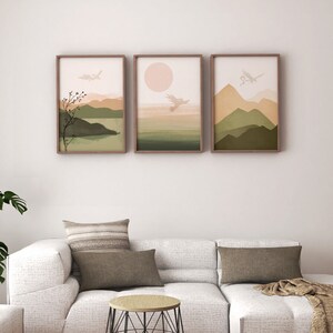 May include: Three framed abstract landscape prints with a muted color palette of green, brown, and pink. The prints feature stylized mountains, a sun, and birds. The prints are hanging on a white wall above a white couch with brown throw pillows.