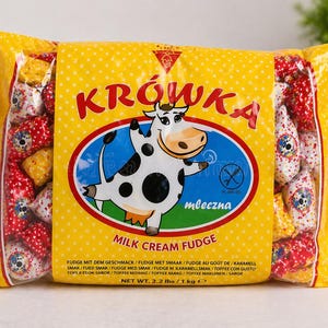 May include: A yellow plastic bag of Krówka milk cream fudge sweets. The packaging features a cartoon cow illustration, the text Krówka and Milk Cream Fudge, and a net weight of 1 kg. The individual sweets inside are wrapped in red, yellow, and white.