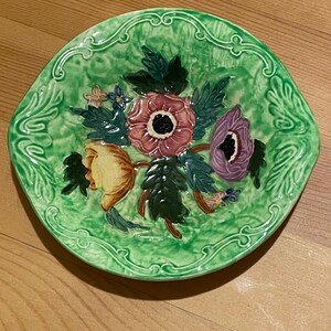 May include: A round ceramic plate with a vibrant green glazed finish and a raised relief floral design. The centre features a cluster of sculpted flowers in shades of pink, yellow, and purple, surrounded by dark green leaves on a textured green background.