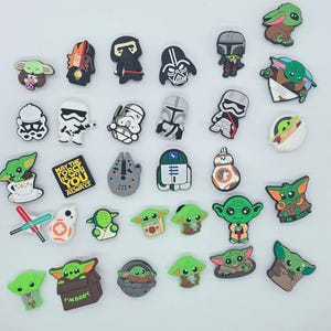 Könnte beinhalten: Eine Sammlung bunter Schuhanhänger mit Charakteren aus dem Star Wars-Franchise. Die Anhänger zeigen Darth Vader, Sturmtruppen, Baby Yoda und andere ikonische Figuren. Die Anhänger bestehen aus einem gummiartigen Material und sind zum Anbringen an Schuhen bestimmt.