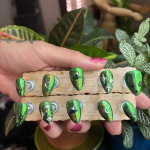 May include: A set of ten press-on nails with a metallic green finish, featuring hand-painted designs including silver crescent moons, starbursts, and alien faces with black eyes. The nails are arranged on two horizontal wooden display stands.