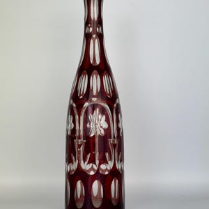 May include: A tall, ruby red glass vase with a dark rim. The vase features a cut glass design with white oval and floral patterns. The elegant shape and color make it a decorative piece for any home.