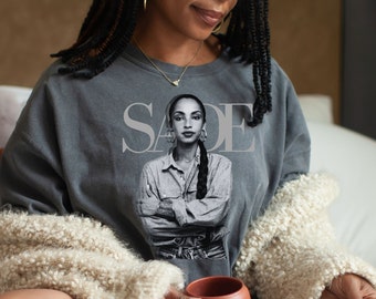 May include: A gray sweatshirt with a black and white image of Sade on the front. The text "SADE" is printed in white above the image.