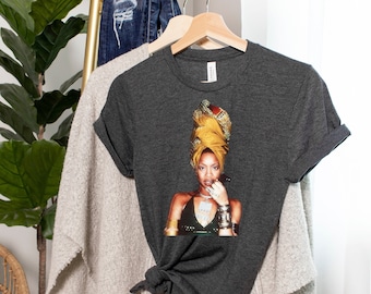 May include: A dark gray t-shirt with a printed image of a woman wearing a yellow head wrap and a silver necklace. The woman is looking at the camera and has her finger to her lips.