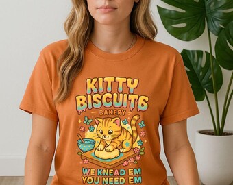 Puede incluir: Camiseta naranja con el texto "KITTY BISCUITS BAKERY" y un gato de dibujos animados amasando masa. El diseño incluye un cuenco, flores y la frase "WE KNEAD EM YOU NEED EM".