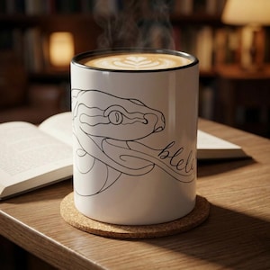 May include: A white ceramic mug with a black rim, featuring a black line art illustration of a snake head and the cursive text blelele. The mug is filled with a hot beverage topped with latte art foam and sits on a round cork coaster.