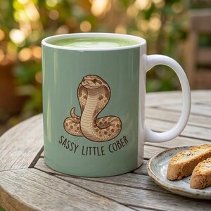 May include: A sage green ceramic mug featuring a brown cobra illustration centred on the front. Below the snake, the text reads SASSY LITTLE COBER in dark brown capital letters. The mug has a white interior and a white handle on the right side.
