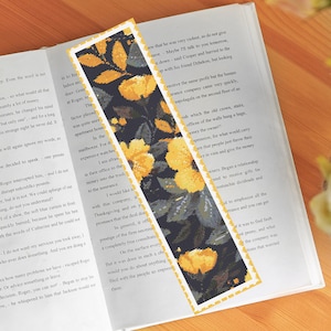 Puede incluir: Un marcapáginas con un diseño floral con flores amarillas y hojas verde oscuro sobre un fondo negro. El marcapáginas tiene un borde dorado.