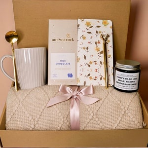 May include: A gift box filled with a cream-colored blanket, a white mug, a gold spoon, a bar of milk chocolate, a floral notebook, a pen, and a candle. The blanket is tied with a pink ribbon. The candle reads "Today's To-Do List: Smile, Be Kind, Spread Happiness."