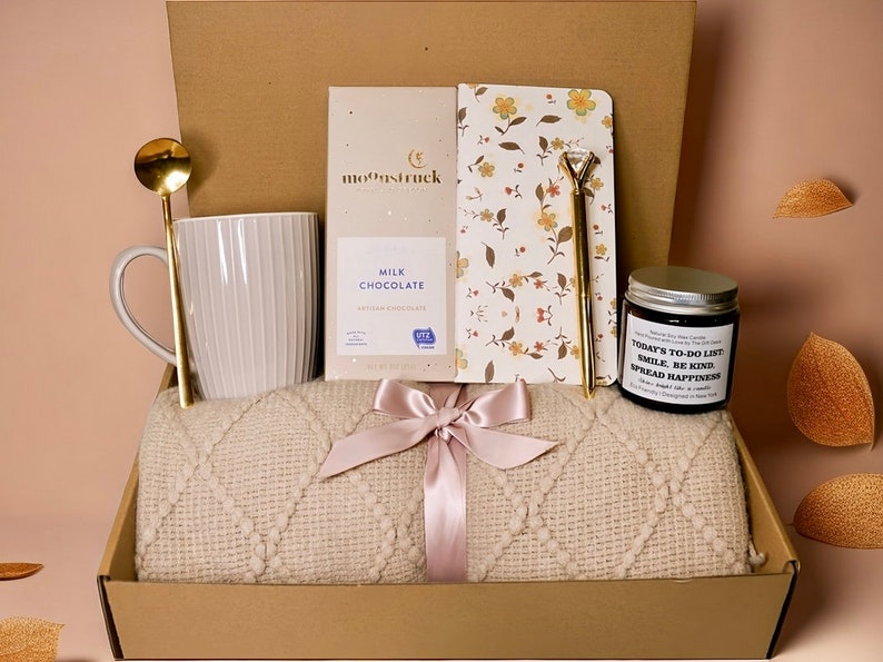May include: A gift box filled with a cream-colored blanket, a white mug, a gold spoon, a bar of milk chocolate, a floral notebook, a pen, and a candle. The blanket is tied with a pink ribbon. The candle reads "Today's To-Do List: Smile, Be Kind, Spread Happiness."
