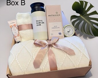May include: A gift box labeled "Box B" containing a white blanket with a pink bow, a travel tumbler, a bar of milk chocolate, a hand cream, a small towel, and a candle. The tumbler reads "TRAVEL TUMBLER 350M."