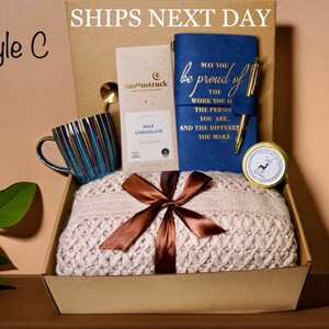 May include: A gift box labeled "Style C" containing a blue and gold striped mug, a bar of milk chocolate, a blue journal with gold text, a gold pen, a candle, and a cream-colored knitted blanket tied with a brown ribbon.