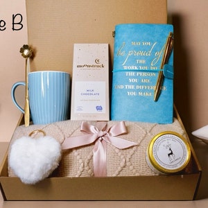 May include: A gift box labeled "Style B" containing a light blue mug, a gold spoon, a bar of milk chocolate, a blue journal with gold text, a gold pen, a cream-colored blanket, a white heart-shaped keychain, and a candle.