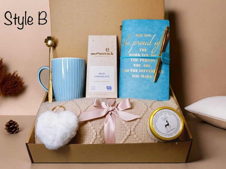 May include: A gift box labeled "Style B" containing a light blue mug, a gold spoon, a bar of milk chocolate, a blue journal with gold text, a gold pen, a cream-colored blanket, a white heart-shaped keychain, and a candle.
