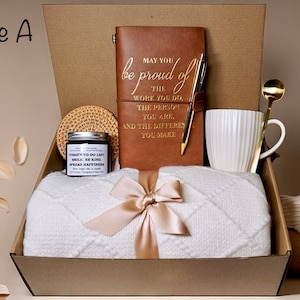 May include: A gift box labeled "Style A" filled with a white blanket, a brown leather journal with gold lettering, a candle, a mug, and a gold spoon. The journal reads "May you be proud of the work you do..."