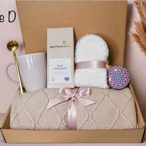 May include: A gift box labeled "Style D" containing a gray mug, gold spoon, chocolate bar, white washcloth, decorative stone, and a beige blanket tied with a pink ribbon. The box is brown.