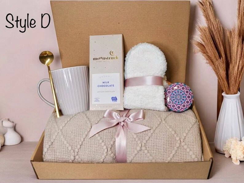 May include: A gift box labeled "Style D" containing a gray mug, gold spoon, chocolate bar, white washcloth, decorative stone, and a beige blanket tied with a pink ribbon. The box is brown.