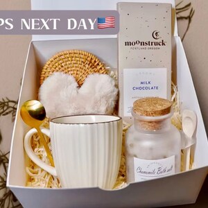 May include: A gift box containing a white mug, gold spoon, heart-shaped eye mask, woven coaster, milk chocolate bar, and a jar of chamomile bath salt. The box is filled with shredded paper.