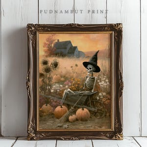 May include: A framed wall art print titled PUDNAMBUT PRINT featuring a skeleton wearing a black witch hat sitting in a wooden wheelbarrow. The scene includes orange pumpkins, sunflowers, and a house in the background under a warm sunset sky.
