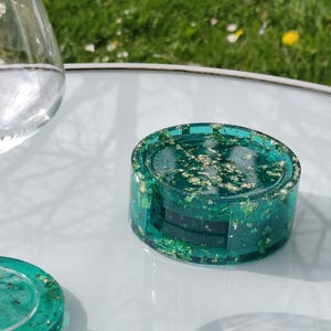 Peut inclure: Un ensemble de quatre dessous de verre en résine turquoise avec des accents de feuilles d'or. Les dessous de verre sont empilés, avec un dessous de verre séparé. Sur une surface blanche, avec un verre et un arrière-plan herbeux.