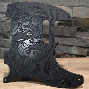 Puede incluir: Un golpeador de guitarra negro con superficie texturizada, un diseño de dragón grabado y caracteres kanji japoneses. El componente incluye orificios para tornillos preperforados y una abertura para la pastilla, con un acabado mate uniforme.