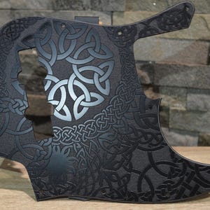 Könnte beinhalten: Ein schwarzes Schlagbrett für E-Bässe mit einem komplexen, geprägten keltischen Knotenmuster auf der gesamten Oberfläche. Das Design zeigt verschiedene ineinandergreifende geometrische Formen und kreisförmige Motive mit matter Oberfläche.