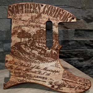 Peut inclure: Un médiator de guitare en bois avec un motif gravé au laser représentant le texte "Southern Comfort" et "The Grand Old Drink of the South". Le motif comprend également une représentation d'une scène de rivière avec des arbres et des bâtiments.