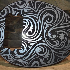 May include: A black and silver bass guitar pickguard featuring a detailed, swirling tribal pattern. The pickguard has a large aperture for the pickup and control knobs, plus several screw holes for mounting. The design is etched into the black surface, creating a striking contrast.
