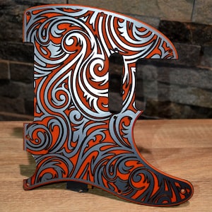 May include: An orange and silver guitar pickguard with a decorative, swirling pattern. The pickguard has a black outline and a cutout for the pickup. The design is symmetrical and detailed, with a mix of curved and straight lines.