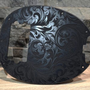 Puede incluir: Golpeador de guitarra negro con un intrincado diseño floral. El golpeador tiene una forma única con un recorte para las pastillas y los controles. La superficie es negro mate y el patrón floral está grabado en el material.