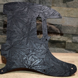 Może przedstawiać: Czarny pickguard do gitary z szczegółowym wzorem liści. Pickguard ma unikalny kształt z kilkoma otworami na śruby i wycięciem na przetwornik. Wzór liści jest wytrawiony na powierzchni, tworząc teksturowany wygląd.