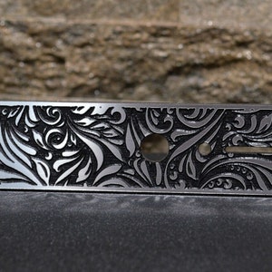 Puede incluir: Una placa de control de guitarra plateada con un patrón floral negro. La placa tiene tres agujeros para botones y una ranura para un interruptor.