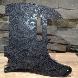 May include: Black guitar pickguard with an intricate paisley design. The pickguard is shaped to fit an electric guitar and features a detailed, engraved pattern across its surface. The design includes floral and swirling elements.