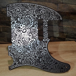May include: A guitar pickguard with an elaborate floral pattern. The pickguard is black with a silver floral design. The pattern includes swirling lines, leaves, and flower shapes. The pickguard has a distinctive shape with a curved base and a cutout for the pickups.