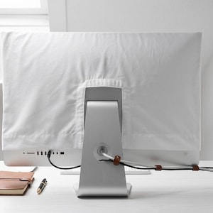 Könnte beinhalten: Eine weiße Monitorabdeckung über einem silbernen Computermonitor. Ein braunes Notizbuch aus Leder, ein Stift und Kabelbinder liegen auf dem weißen Schreibtisch. Der Monitor steht auf einem weißen Schreibtisch in einem hellen Raum.