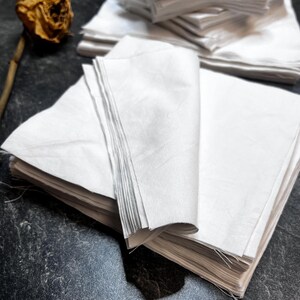 Puede incluir: Una pila de piezas de tela blanca cuadradas con bordes sin rematar, dispuestas en un montón ordenado con una esquina doblada. Una rosa marrón seca y marchita con un tallo largo se sitúa a la izquierda de la pila de tela sobre una superficie oscura.