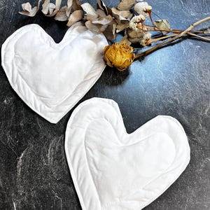 Può includere: Due cuscini in tessuto bianco a forma di cuore con bordi cuciti. I cuscini presentano una consistenza morbida e imbottita con una superficie bianca liscia. Sono disposti accanto a un rametto di foglie di eucalipto essiccate e un fiore giallo secco.